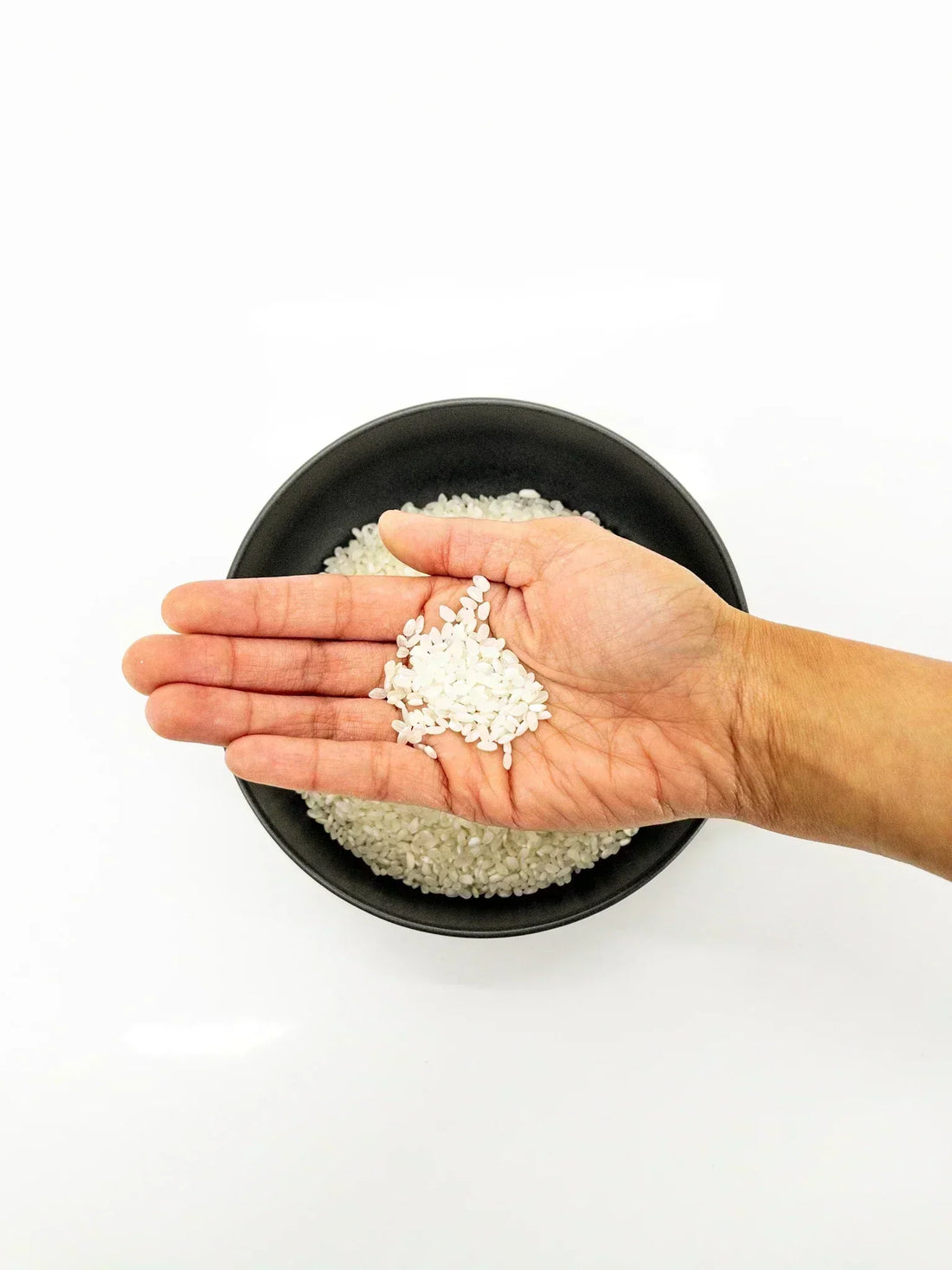 À l'avant plan, une main tenant des grains de riz secs, à l'arrière plan, un bol noir contenant d'autres grains de riz secs | Eat in Japan épicerie en ligne